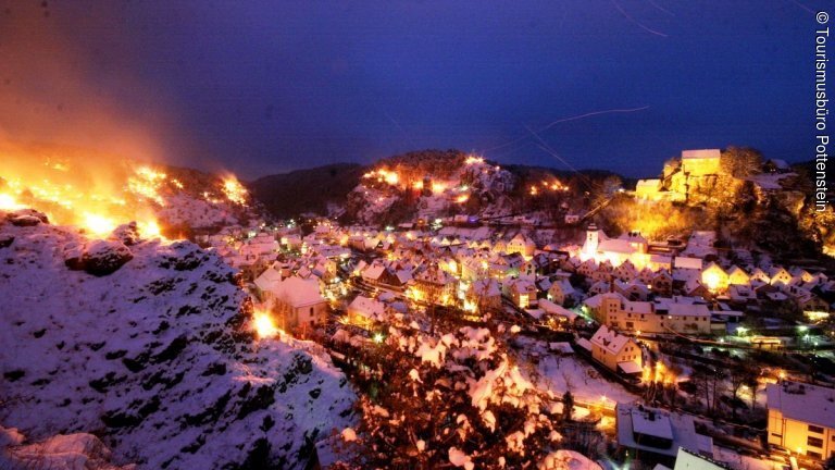 Nächtlicher Blick auf die Felsenstadt Pottenstein mit beleuchteten Häusern und Feuern an den Berghängen.