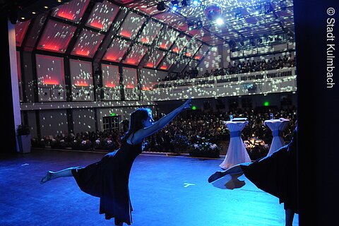 Tänzerin in schwarzem Kleid auf Bühne vor Publikum mit Disco-Lichteffekt an Decke und Wänden