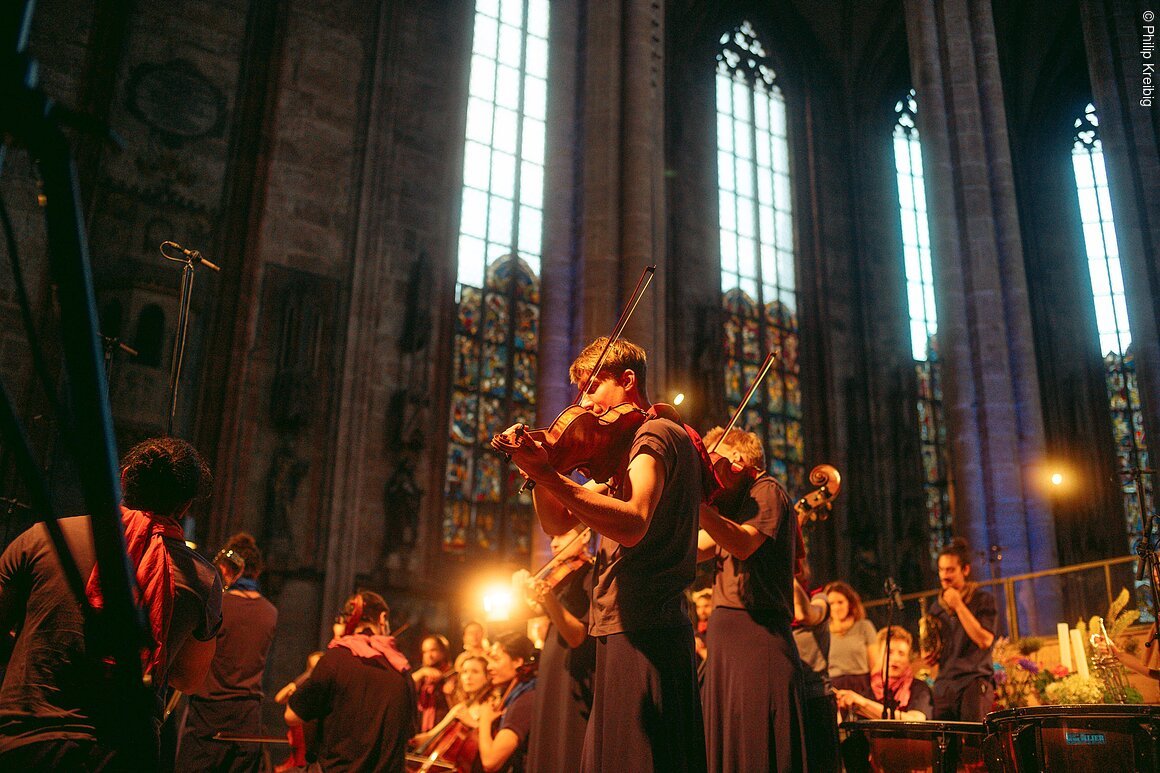 Junge Musiker spielen Streichinstrumente in einer Kirche mit hohen Fenstern und bunten Glasmalereien.