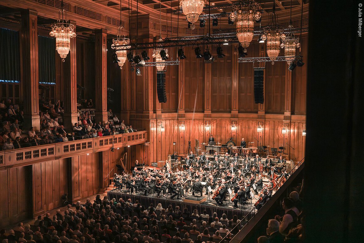 Orchester spielt in einem Konzertsaal mit Holzverkleidung vor großem Publikum und Kronleuchtern.