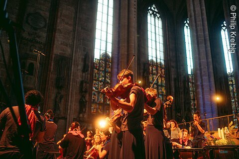 Junge Musiker spielen Streichinstrumente in einer Kirche mit hohen Fenstern und bunten Glasmalereien.