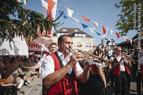 Mann in roter Weste spielt Trompete bei Straßenfest mit blau-weiß-roten Wimpeln und Publikum im Hintergrund.