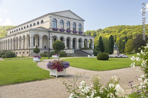 Historisches Gebäude mit Arkaden und Blumen in weißen Kästen in einem gepflegten Park bei Sonnenschein.