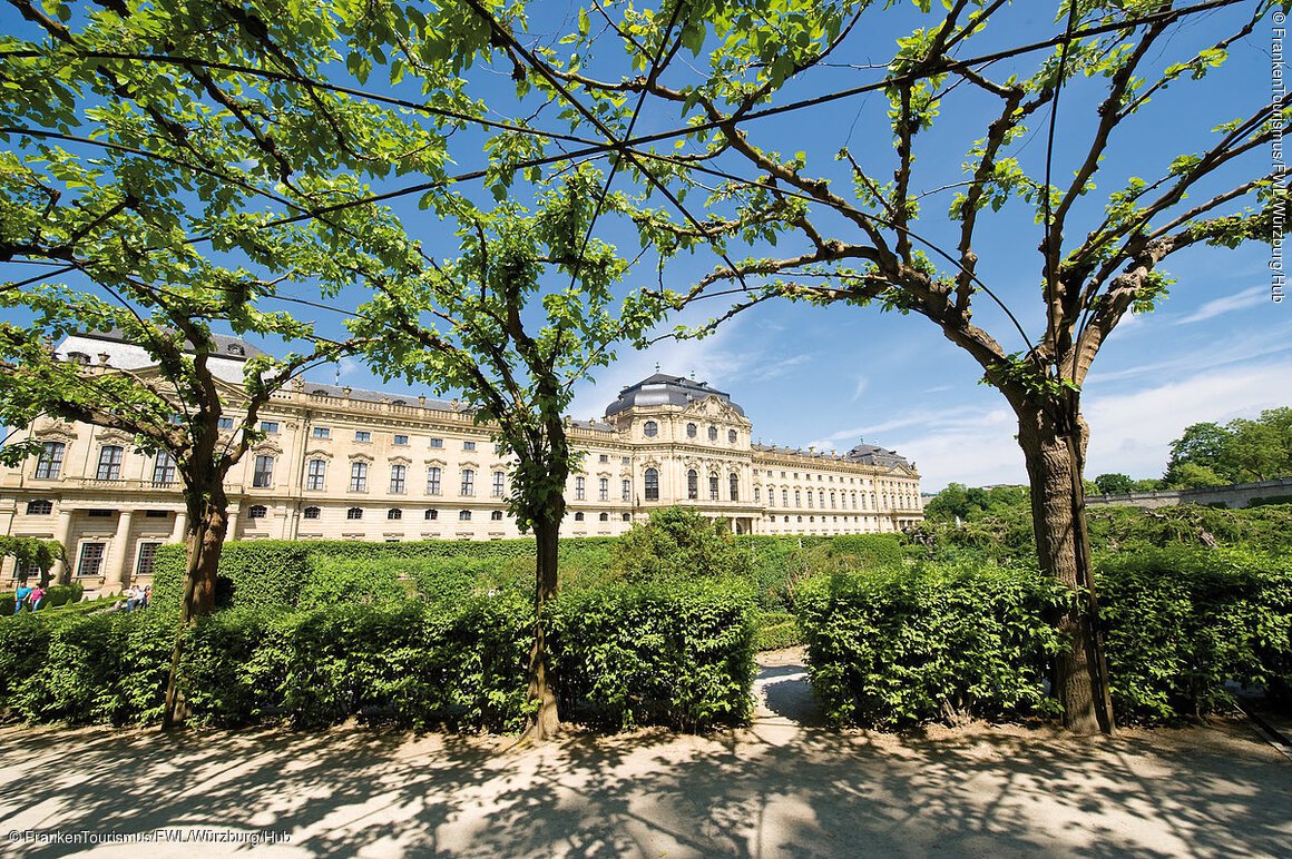 Blick durch grüne Baumreihen auf ein historisches Schlossgebäude bei blauem Himmel.