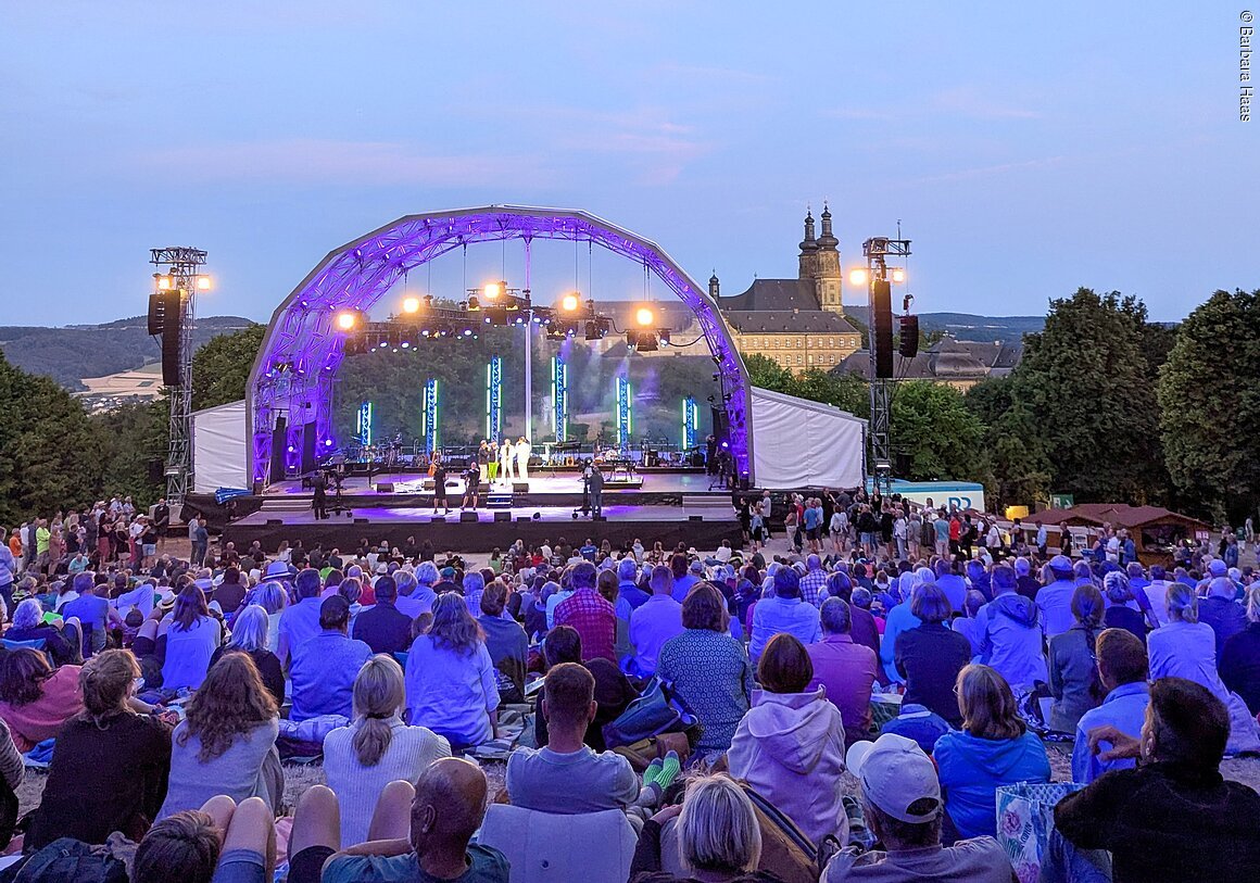 Open-Air-Konzert mit Bühne, Publikum auf Decken sitzend, im Hintergrund ein großes Gebäude mit Türmen bei Abenddämmerung.