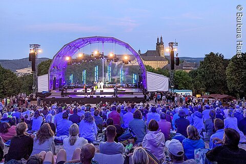 Open-Air-Konzert mit Bühne, Publikum auf Decken sitzend, im Hintergrund ein großes Gebäude mit Türmen bei Abenddämmerung.
