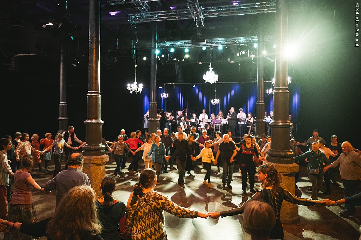 Menschen tanzen im Kreis in einem Saal mit Bühne und Live-Musik im Hintergrund unter Kronleuchtern.