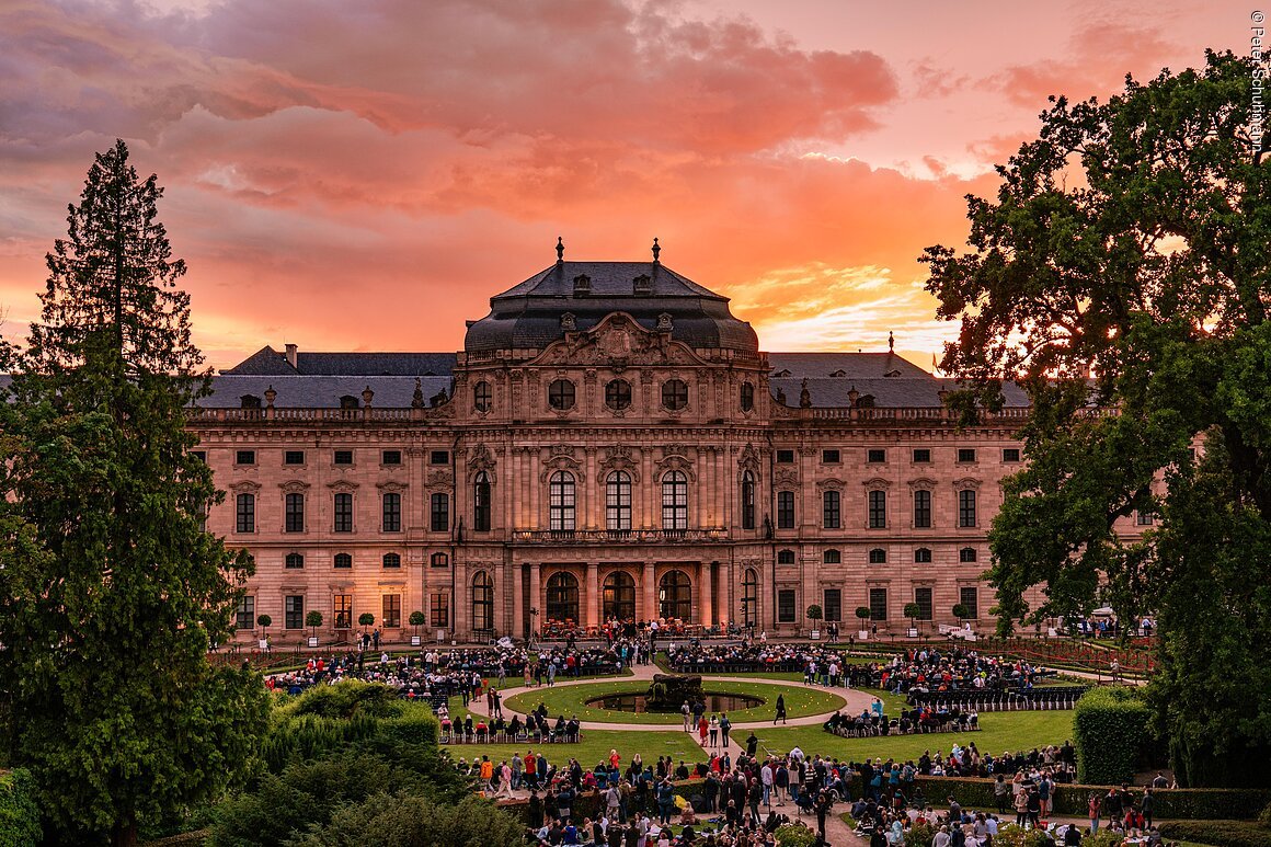 Menschenmenge vor großem historischem Gebäude bei Sonnenuntergang mit orangefarbenem Himmel und Bäumen.