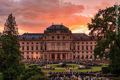 Menschenmenge vor großem historischem Gebäude bei Sonnenuntergang mit orangefarbenem Himmel und Bäumen.
