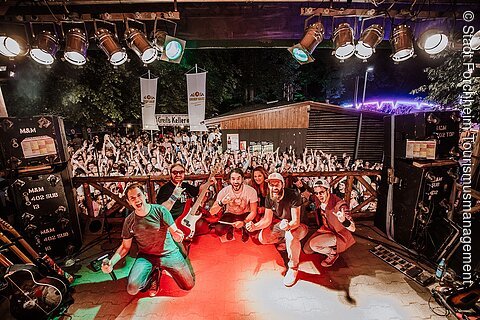 Bandmitglieder posieren vor jubelndem Publikum bei Konzert auf Bühne mit Scheinwerfern und Musikinstrumenten.