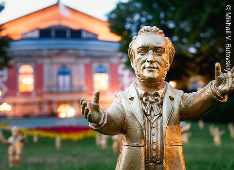 Goldfarbene Statue eines Mannes mit ausgestreckten Armen vor einem beleuchteten historischen Gebäude bei Dämmerung