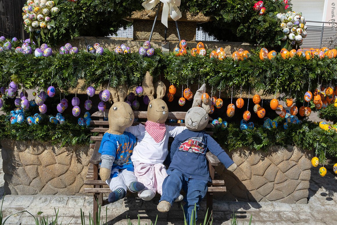 Drei Hasenfiguren aus Stoff sitzen auf einer Bank vor einem mit bunten Ostereiern geschmückten grünen Kranz.