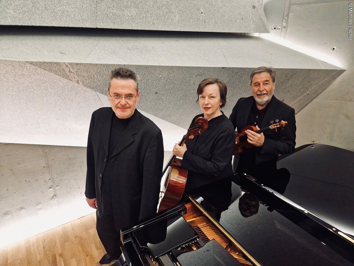 Eine Frau mit Violoncello und zwei Männer, einer davon mit Geige, stehen in einem Saal neben einem Flügel.