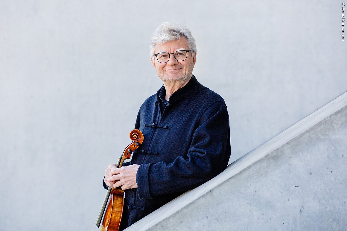 Ein älterer Mann steht hinter einer schräg verlaufenden, halbhohen Betonwand. Er stützt sich mit einem Arm darauf ab und hält eine Viola vor dem Oberkörper.