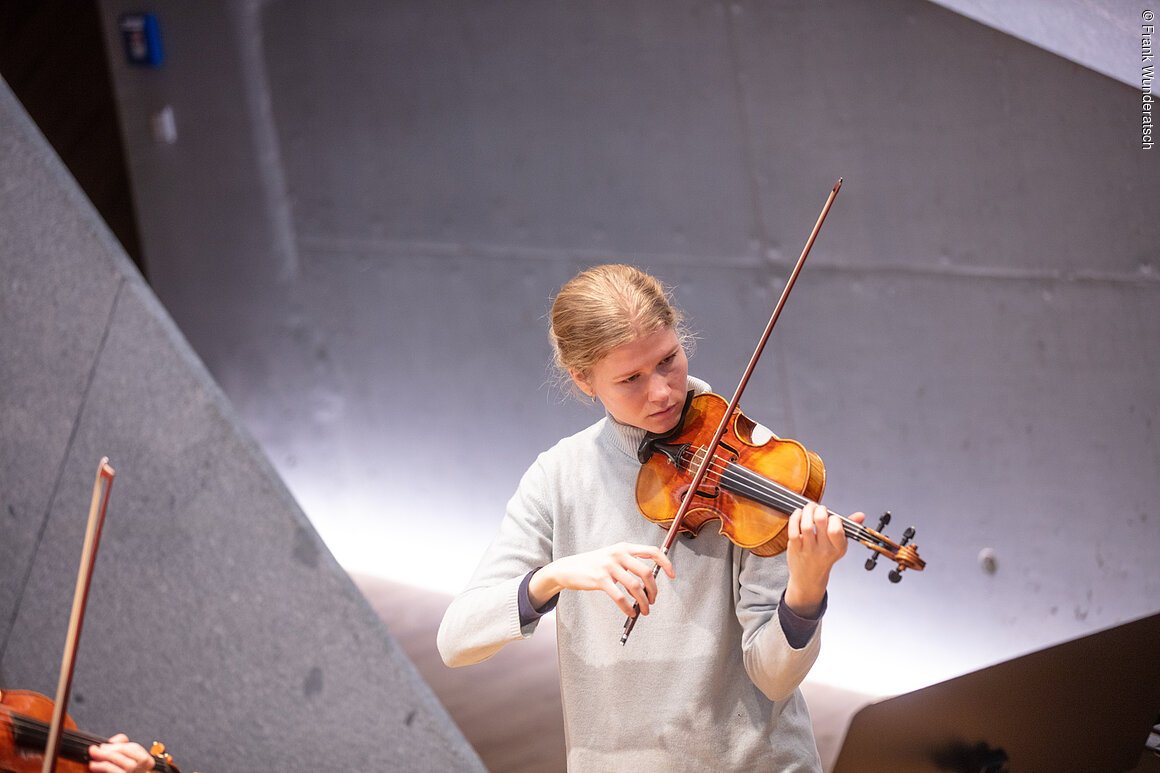 Eine junge Frau im grauen Rollkragenpullover spielt Geige. Im Raumhintergrund sind geometrische Formen mit effektvoller Beleuchtung zu erkennen. Am linken Bildrand sind Teile einer Geige und eines Bogens zu sehen.