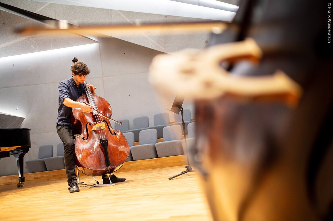 Eine Person mit einem Kontrabass steht mit seinem Instrument in einem Raum mit Gesteinselementen. Er setzt den Bogen an..Am, rechten Bildrand ist unscharf der Steg eiees weiteren Kontrabasses zu sehen, links ein Teil eines Flügels.
