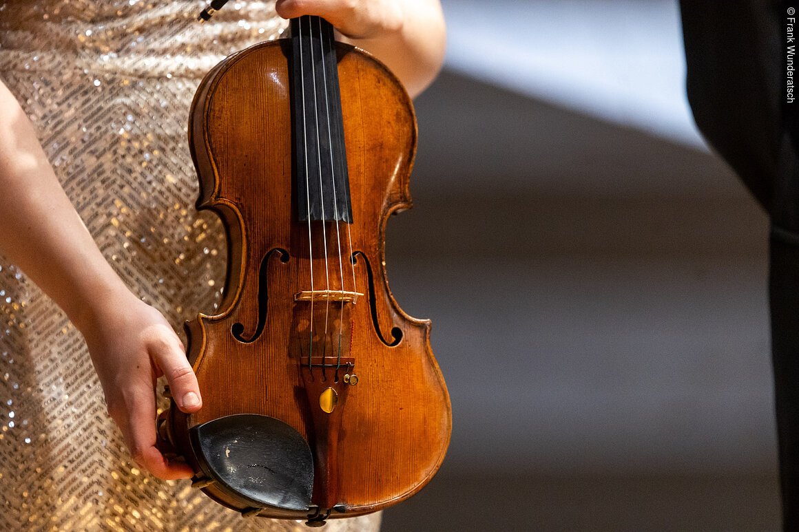 Eine Hand  hält eine Violine, hochkant. Der Hals des Instruments ist nicht zu sehen.