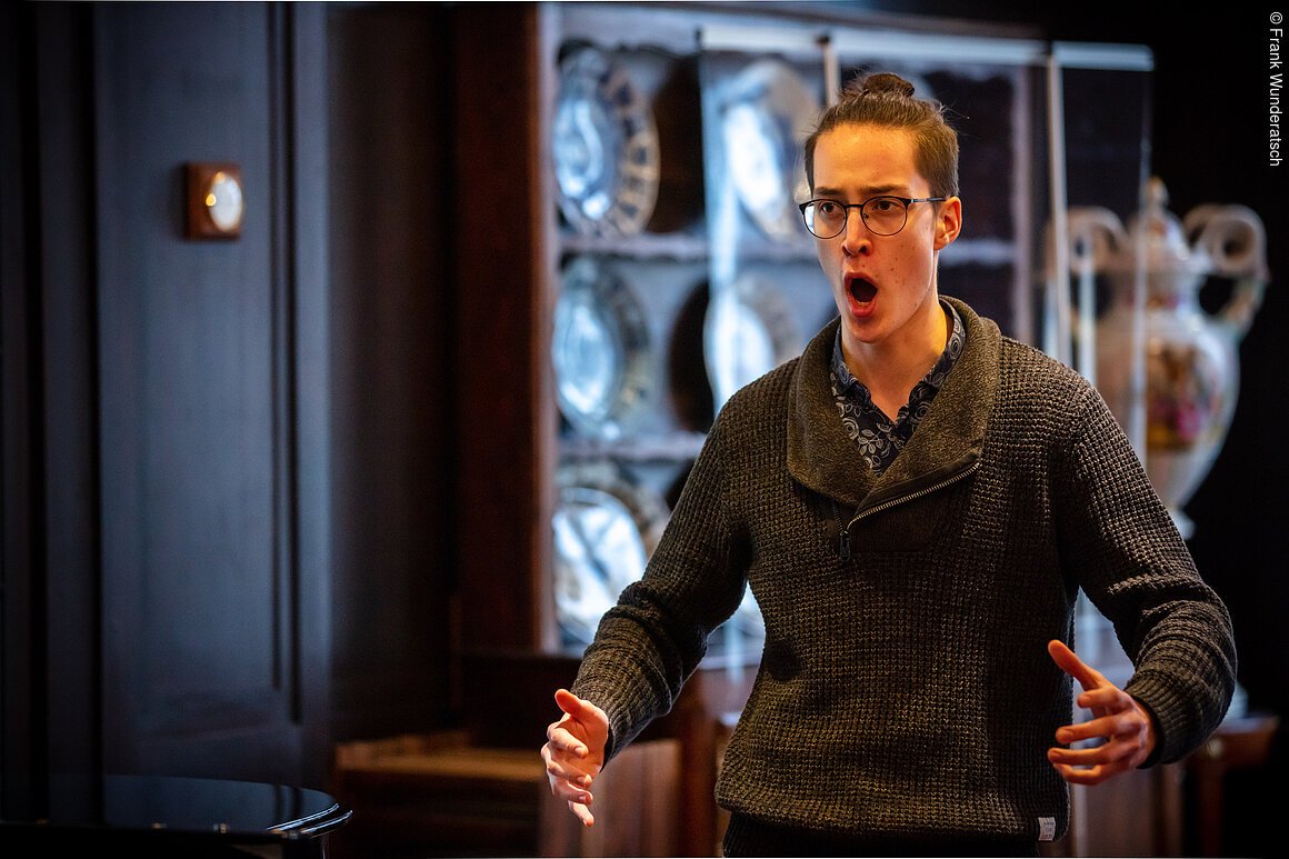 Ein junger Mann singt in einem holzvertäfelten Raum. Im Hintergrund ist eine antike Porzellanvitrine zu sehen.