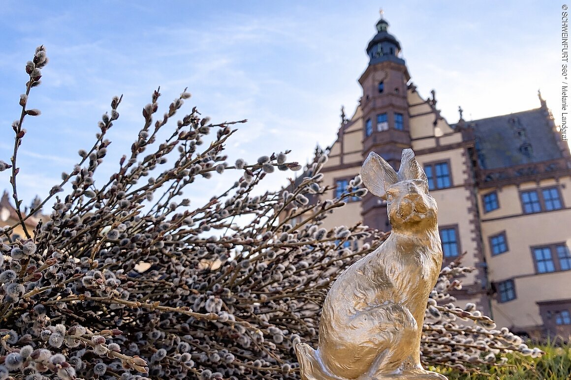 Goldene Hasenskulptur vor einem großen Gebäude und Weidenkätzchen bei bewölktem Himmel