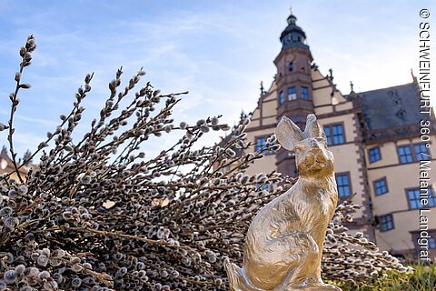 Goldene Hasenskulptur vor einem großen Gebäude und Weidenkätzchen bei bewölktem Himmel