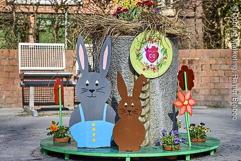 Osterdekoration mit zwei Hasenfiguren, Blumen und einem Brunnen in einem öffentlichen Bereich.