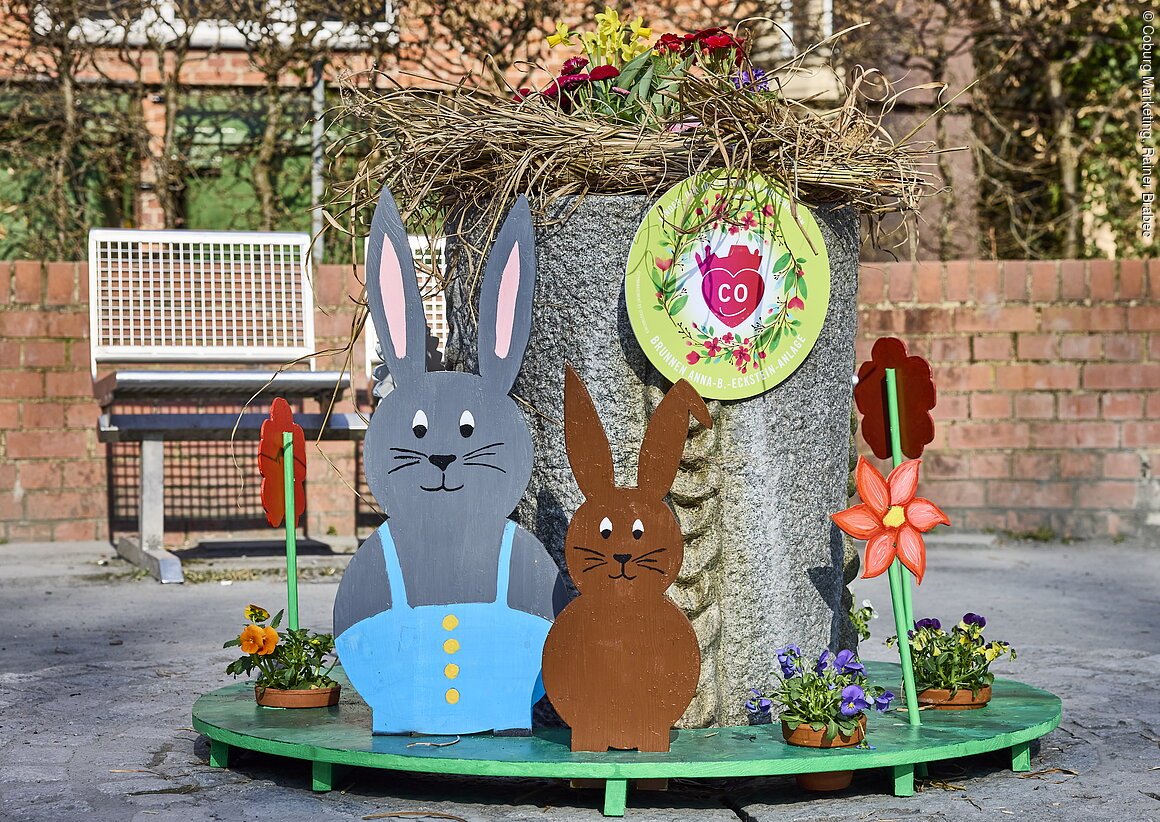 Osterdekoration mit zwei Hasenfiguren, Blumen und einem Brunnen in einem öffentlichen Bereich.