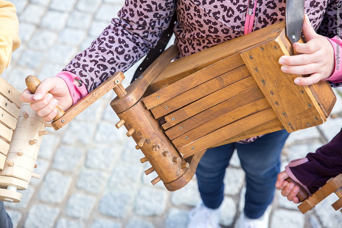 Person hält eine historische Drehleier aus Holz mit Griff und Stimmnägeln, Pflastersteinboden im Hintergrund.