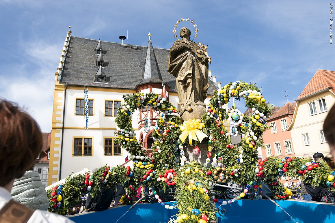 Osterbrunnen mit geschmücktem Kranz aus Tannengrün und bunten Ostereiern vor historischem Gebäude und blauem Himmel.
