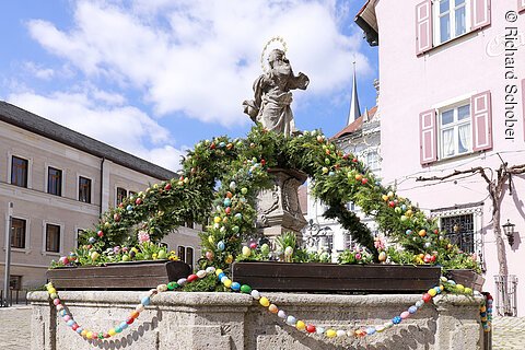 Brunnen mit Statue und geschmückten grünen Bögen sowie bunten Ostereiern in einem Stadtplatz bei Sonnenschein