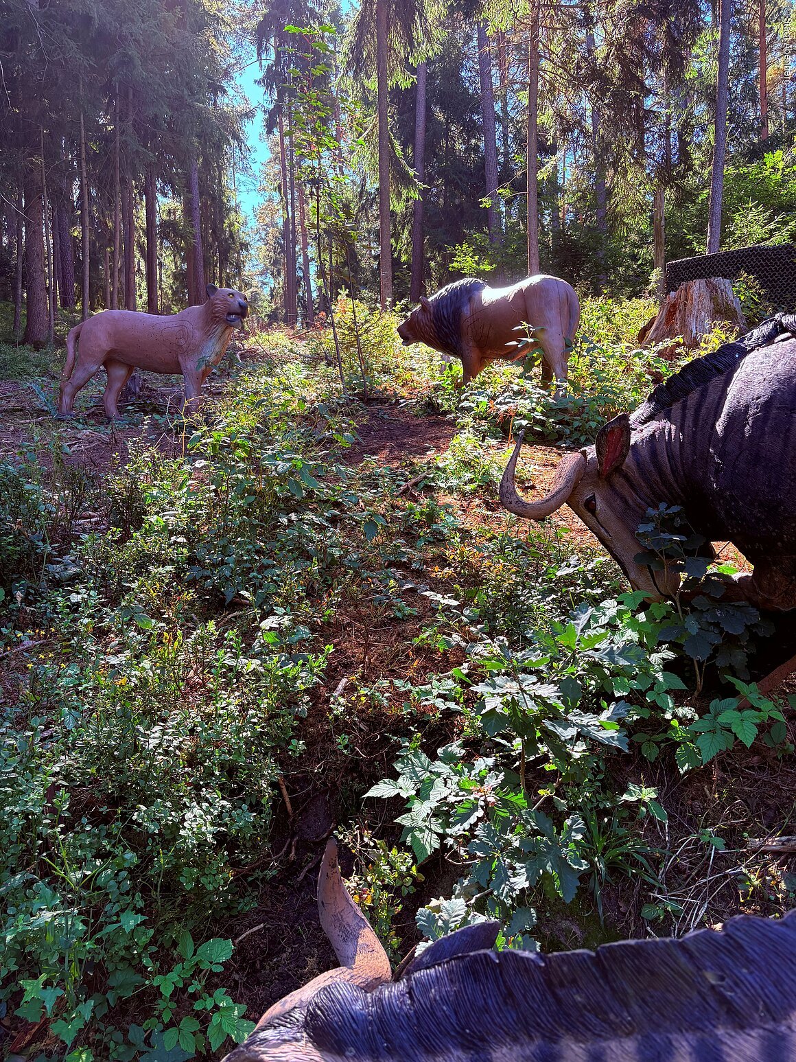 Wald mit drei Tierfiguren aus Holz, darunter ein Löwe, ein Büffel und ein weiteres Tier, umgeben von Pflanzen.