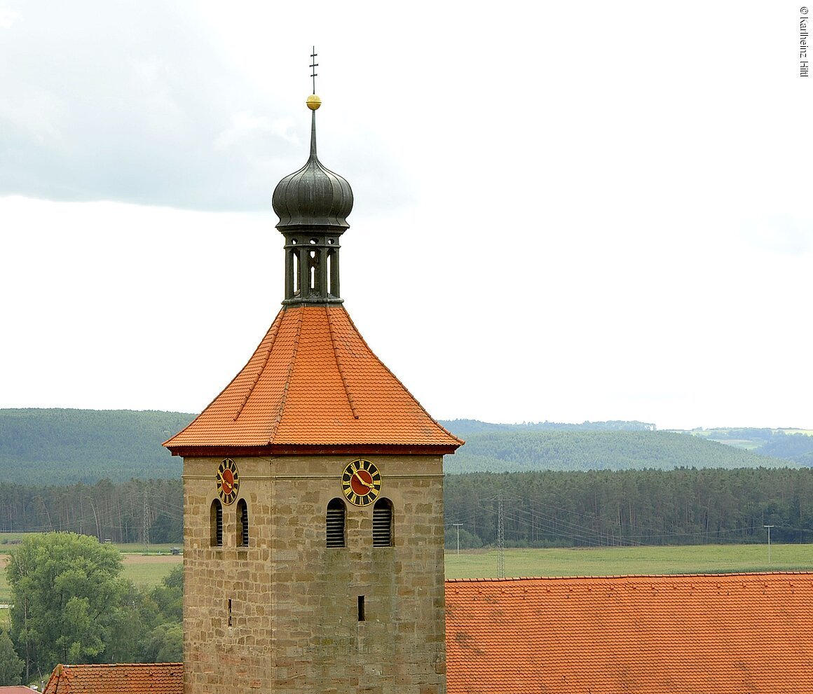 Kirchturm mit rotem Ziegeldach, zwei Uhren und Zwiebelhaube vor bewaldeter Hügellandschaft unter bewölktem Himmel.
