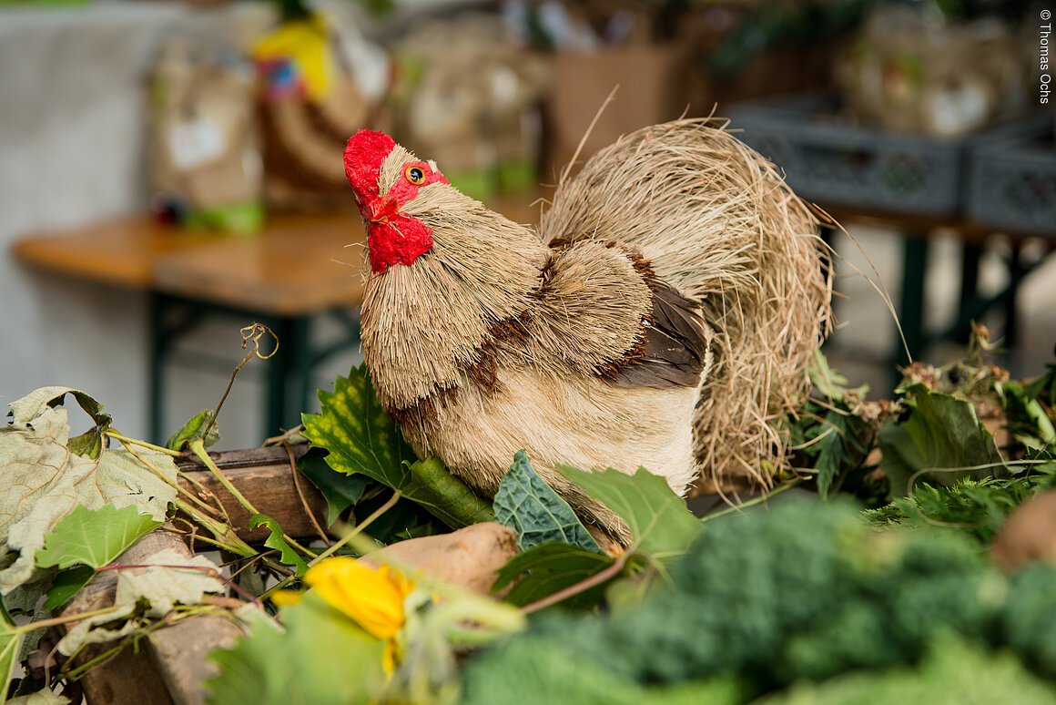 Deko-Huhn aus Stroh mit rotem Kamm steht auf einem Holzkasten mit grünen Blättern und Gemüse.