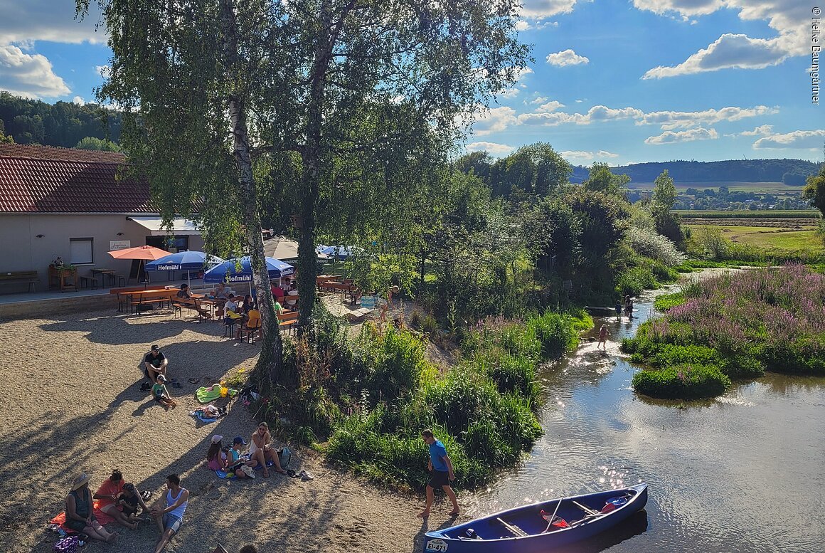 Menschen sitzen am Ufer eines Flusses mit Kanus und Bänken unter Bäumen an einem sonnigen Tag.