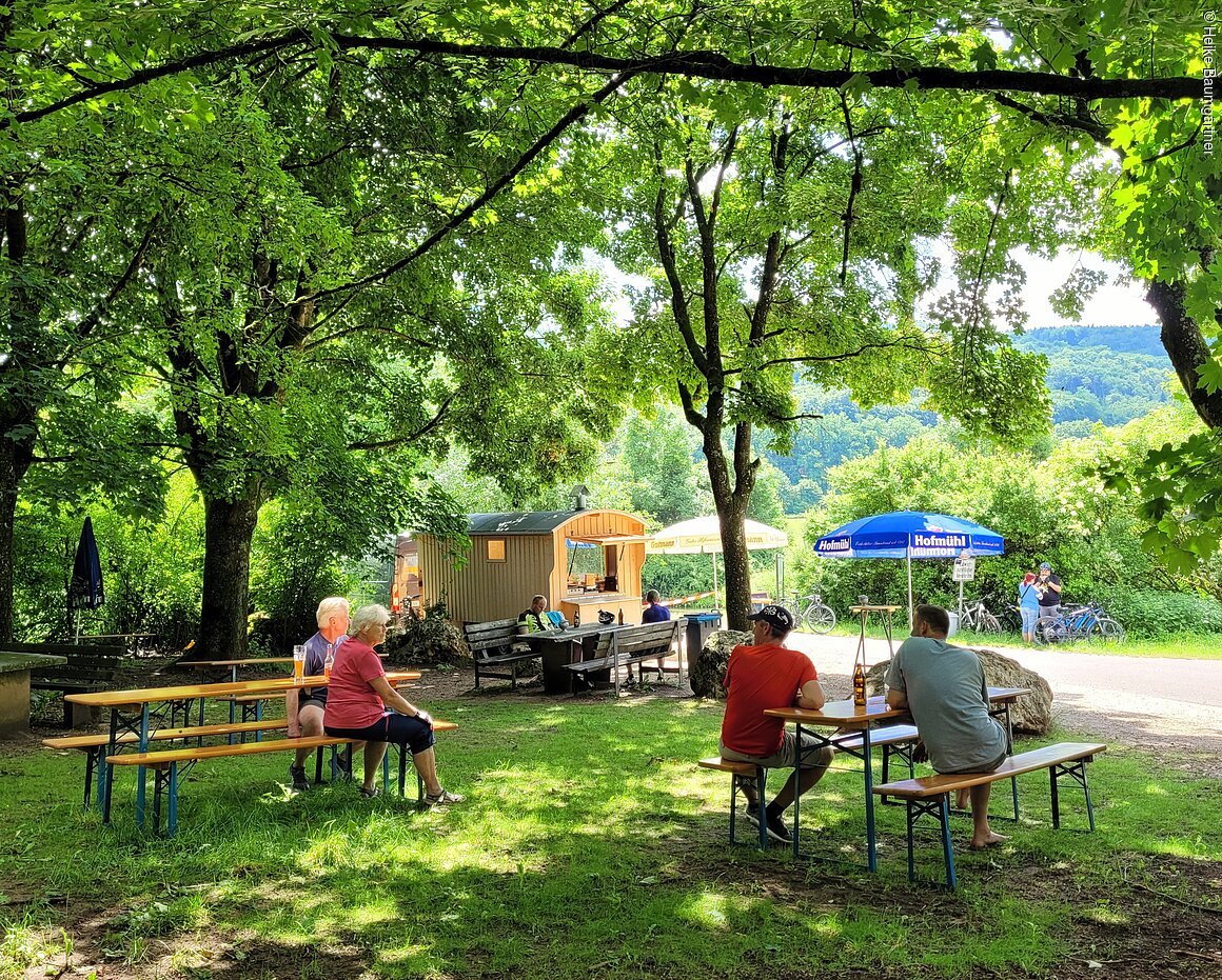 Menschen sitzen an Biergartenbänken unter Bäumen mit Imbisswagen und Sonnenschirmen im Hintergrund.