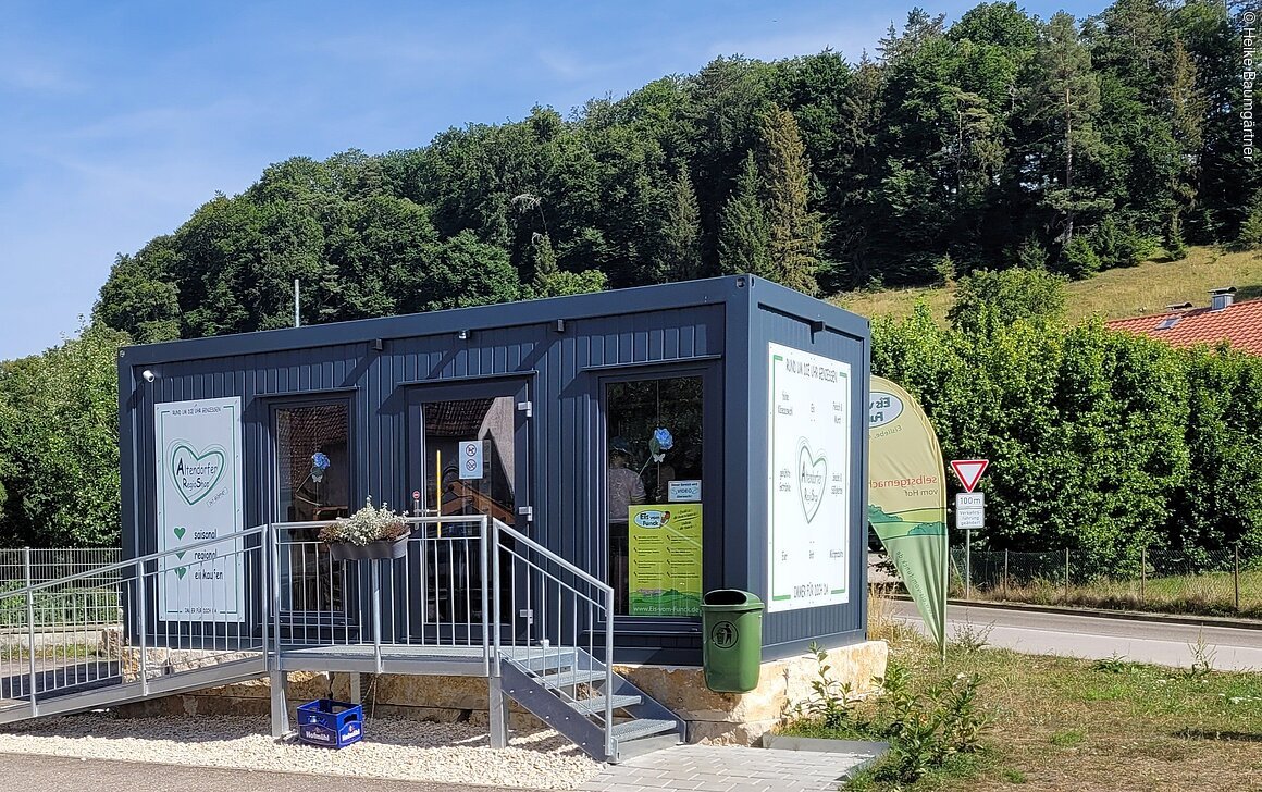 Kleines Verkaufsgebäude mit Rampe, umgeben von Grün und Wald im Hintergrund.