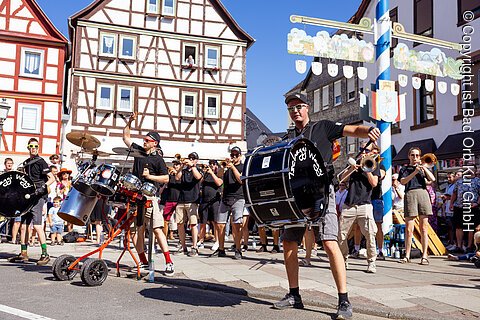 Straßenmusikanten mit Trommeln und Blasinstrumenten vor Fachwerkhäusern bei sonnigem Wetter.