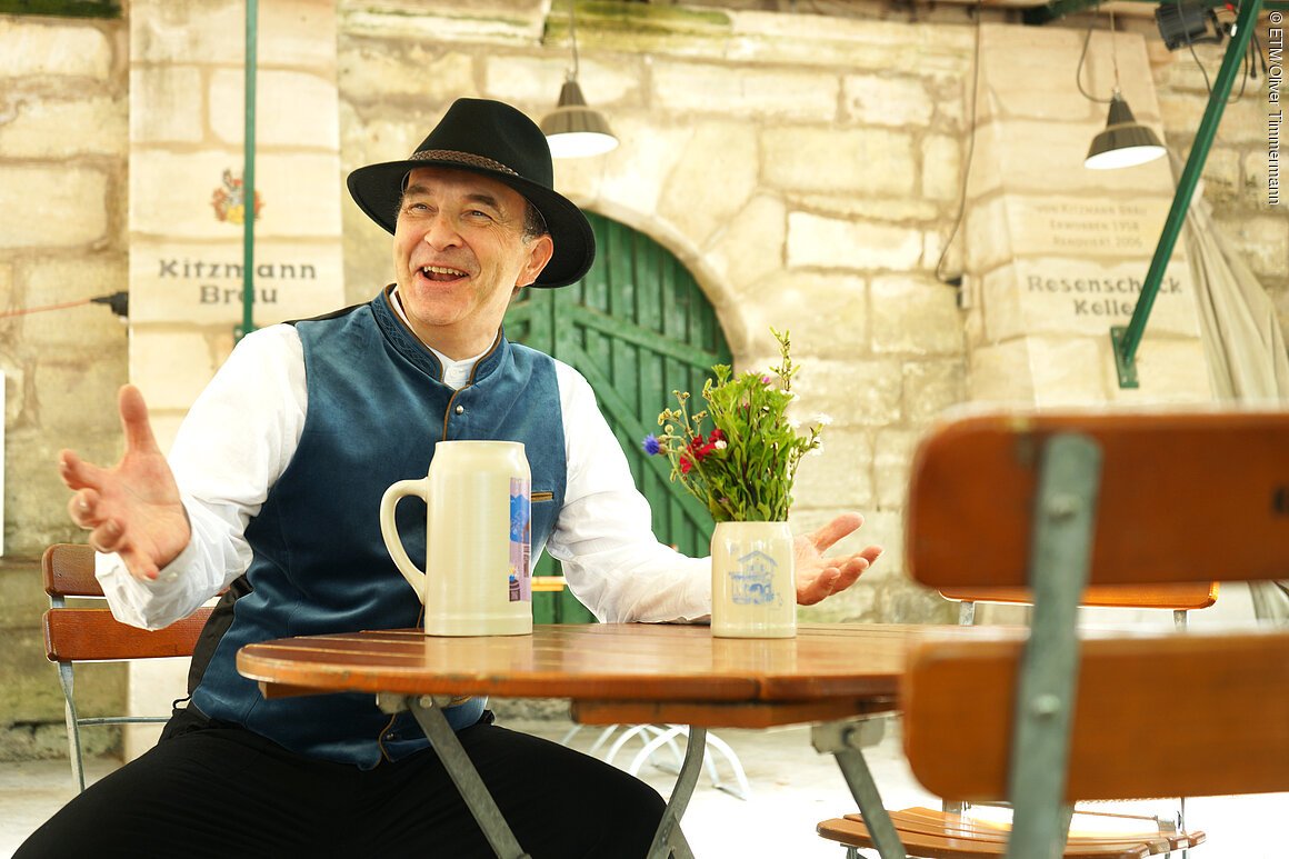 Bierkutscher Schorsch in Trachtenweste und Hut sitzt an Holztisch mit Bierkrug und Blumenvase vor Steinmauer mit grüner Tür.