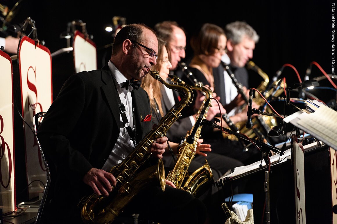 Fünf Musiker in schwarzer Kleidung spielen Saxophone in einer Reihe auf einer Bühne.