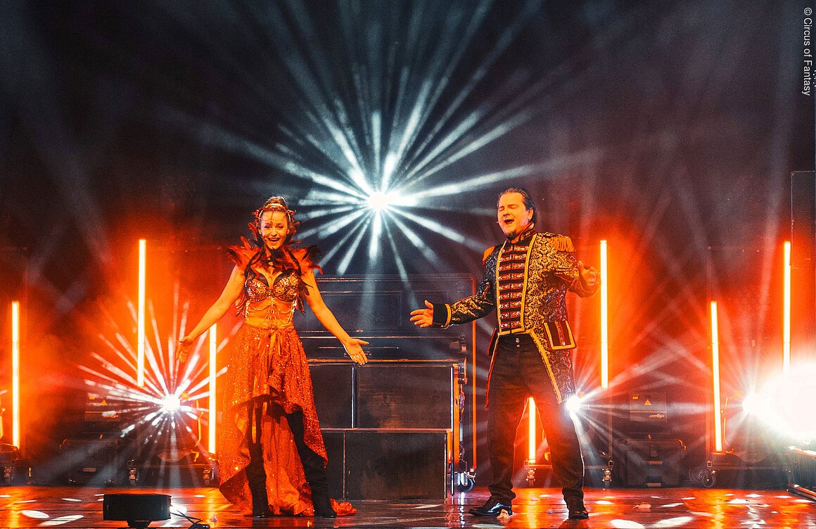Zwei Personen in Bühnenkostümen singen vor leuchtenden Scheinwerfern und orangefarbenem Bühnenlicht.