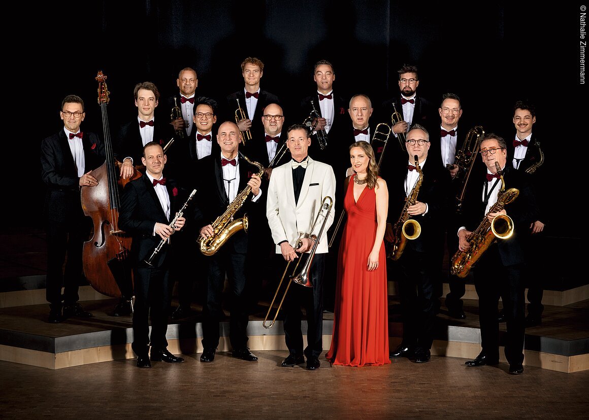 Große Jazzband mit 16 Männern in schwarzen Anzügen und roter Fliege, eine Frau im roten Kleid, Instrumente sichtbar.