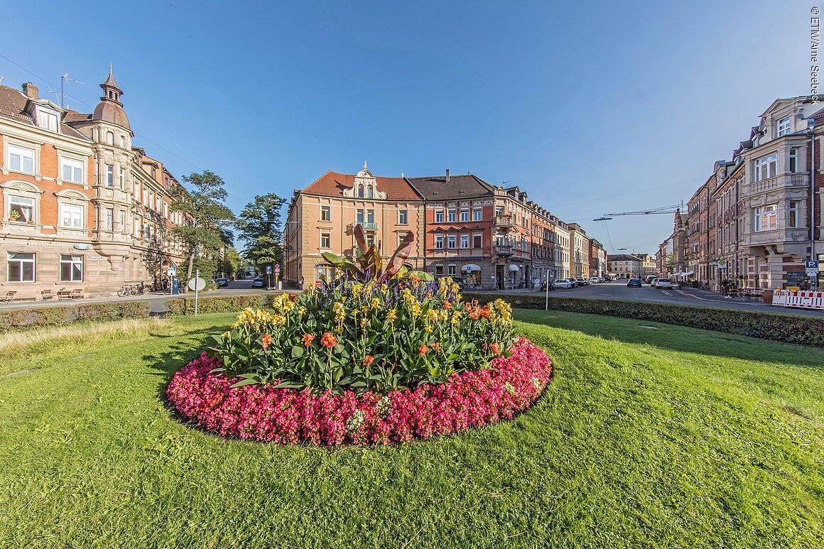 Rundes Blumenbeet mit bunten Blumen auf einer grünen Wiese vor historischen Gebäuden bei klarem Himmel.