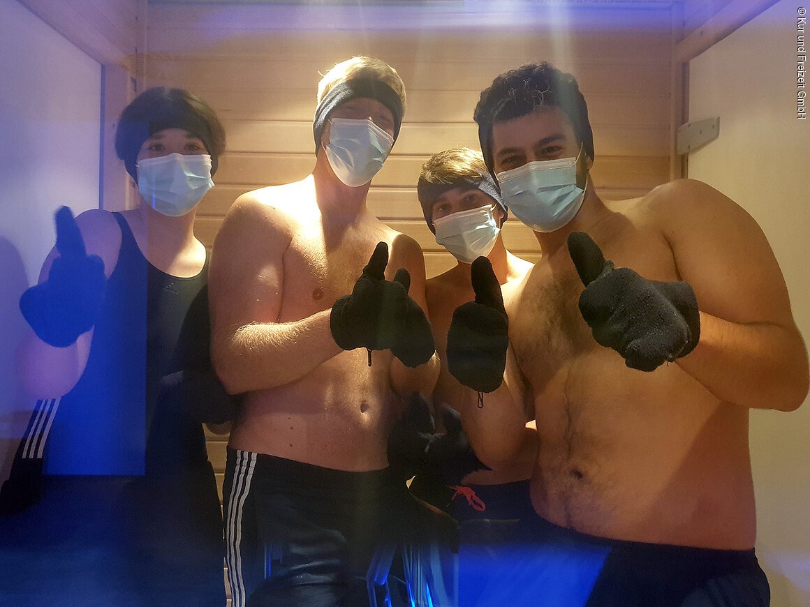 Vier Personen mit Mund-Nasen-Schutz, Stirnbändern und schwarzen Handschuhen zeigen Daumen hoch in einem Holzraum.