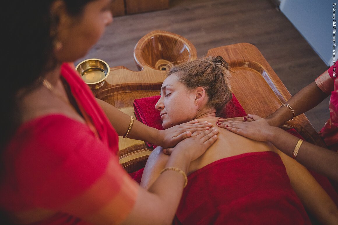Frau liegt mit geschlossenen Augen auf Massagetisch, zwei Personen massieren ihre Schultern.