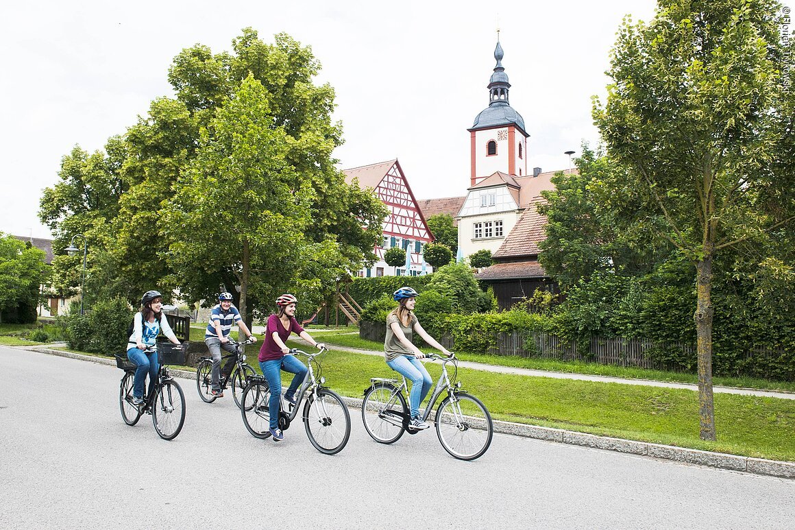 Vier Personen mit Fahrradhelmen fahren auf einer Straße vor Bäumen und einem Fachwerkhaus mit Kirchturm.