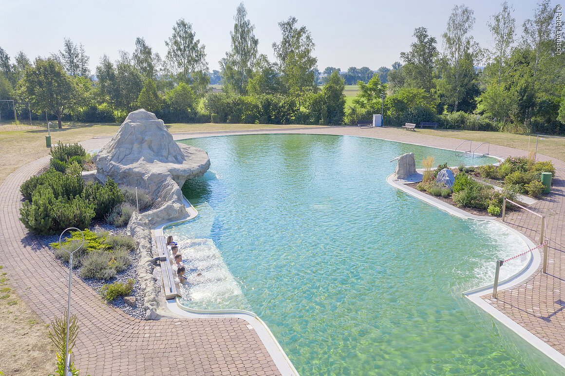 Außenpool mit klar grünem Wasser, Felsenstruktur und vier Personen am Beckenrand sitzend.