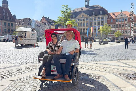 Zwei Personen sitzen in einer roten Rikscha auf einem gepflasterten Marktplatz mit historischen Gebäuden im Hintergrund.
