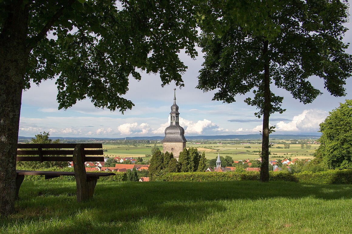 Bank unter Bäumen mit Blick auf Kirchturm und Dorf in ländlicher Umgebung bei Tageslicht