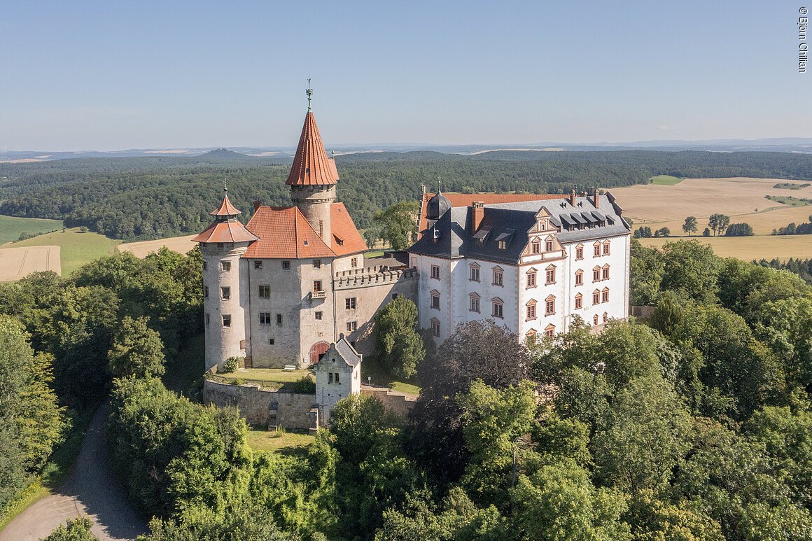 Burg mit roten Dächern umgeben von Bäumen und Feldern unter blauem Himmel, Luftaufnahme.