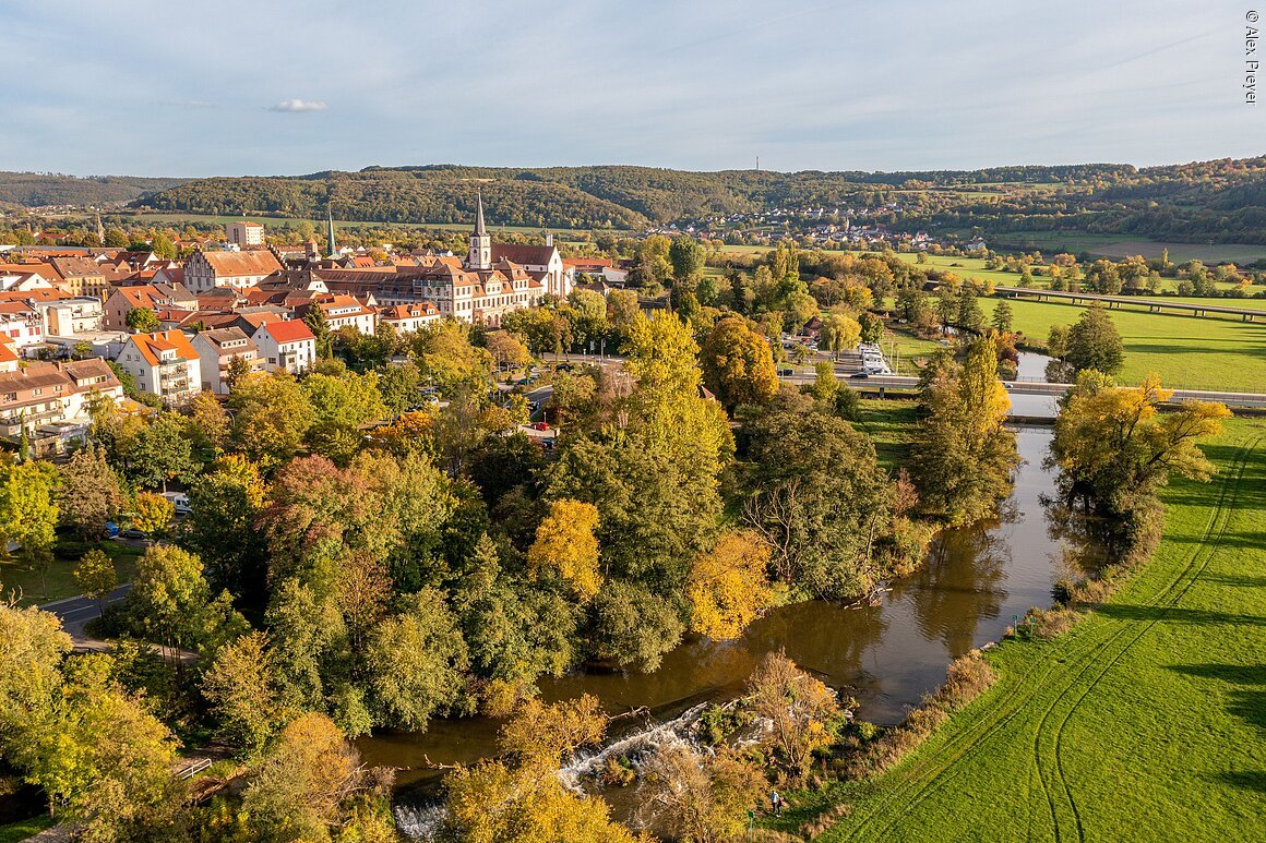 Luftaufnahme einer Stadt mit Fluss, herbstlich gefärbten Bäumen und grünen Wiesen im Umland.