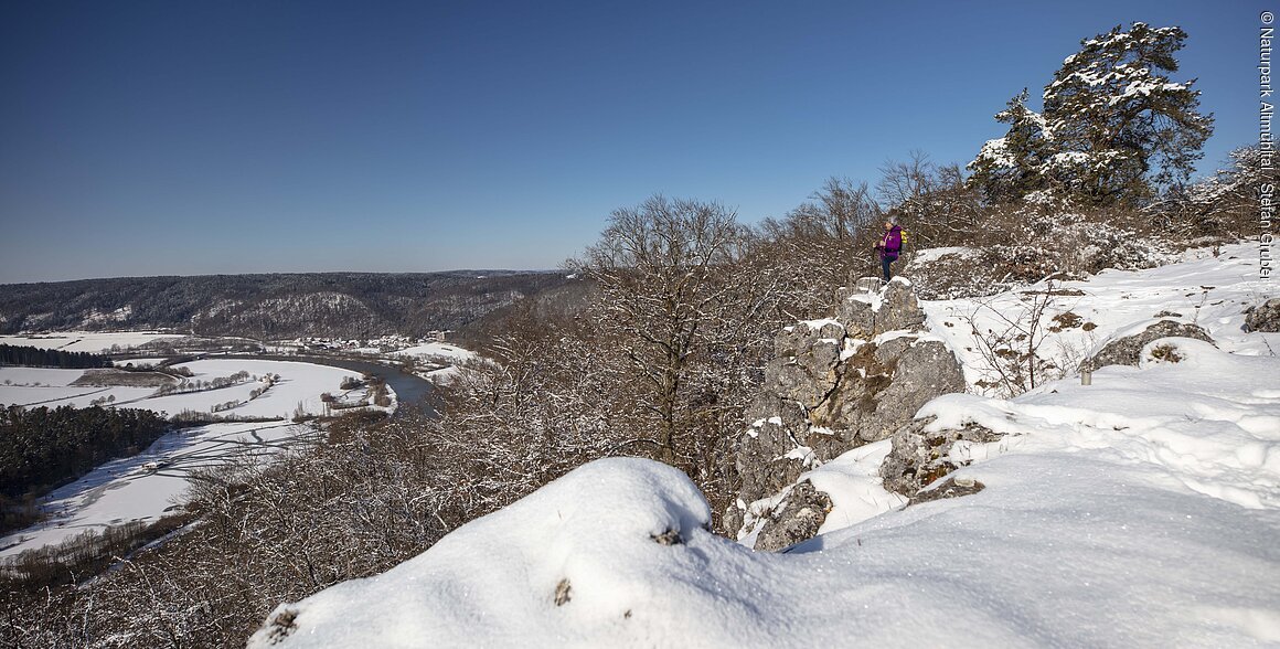 Winterlandschaft mit verschneiten Bäumen, Felsen und einem Fluss im Tal unter blauem Himmel, Wanderer auf Felsen